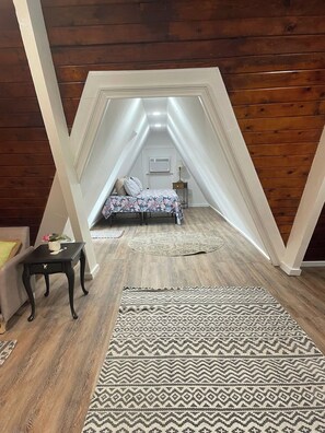 A loft bedroom with a sloped ceiling, a bed with a floral bedspread, a small table with a vase, and a patterned rug. - Poplar Cottage at Saluda River Farms (Greenville)