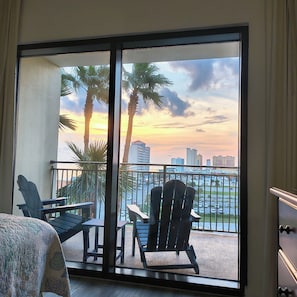 Un balcón con dos sillas Adirondack, vista a palmeras y un paisaje urbano al atardecer.