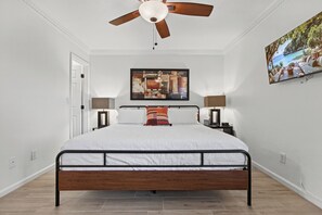 A bedroom with a bed, a ceiling fan, a nightstand with a lamp, a painting on the wall, and a door. - Foothill Garden | Pool, Patio & La Encantada (Tucson)