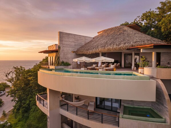 Uma villa moderna com piscina infinita no terraço, uma construção com telhado de palha e vista para o mar.