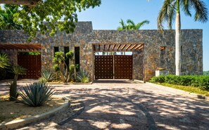 Un edificio de piedra con entrada porticada, palmeras y una pérgola.