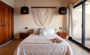 Een slaapkamer met een groot bed, twee nachtkastjes, een macramé wanddecoratie en een groot raam met gordijnen.