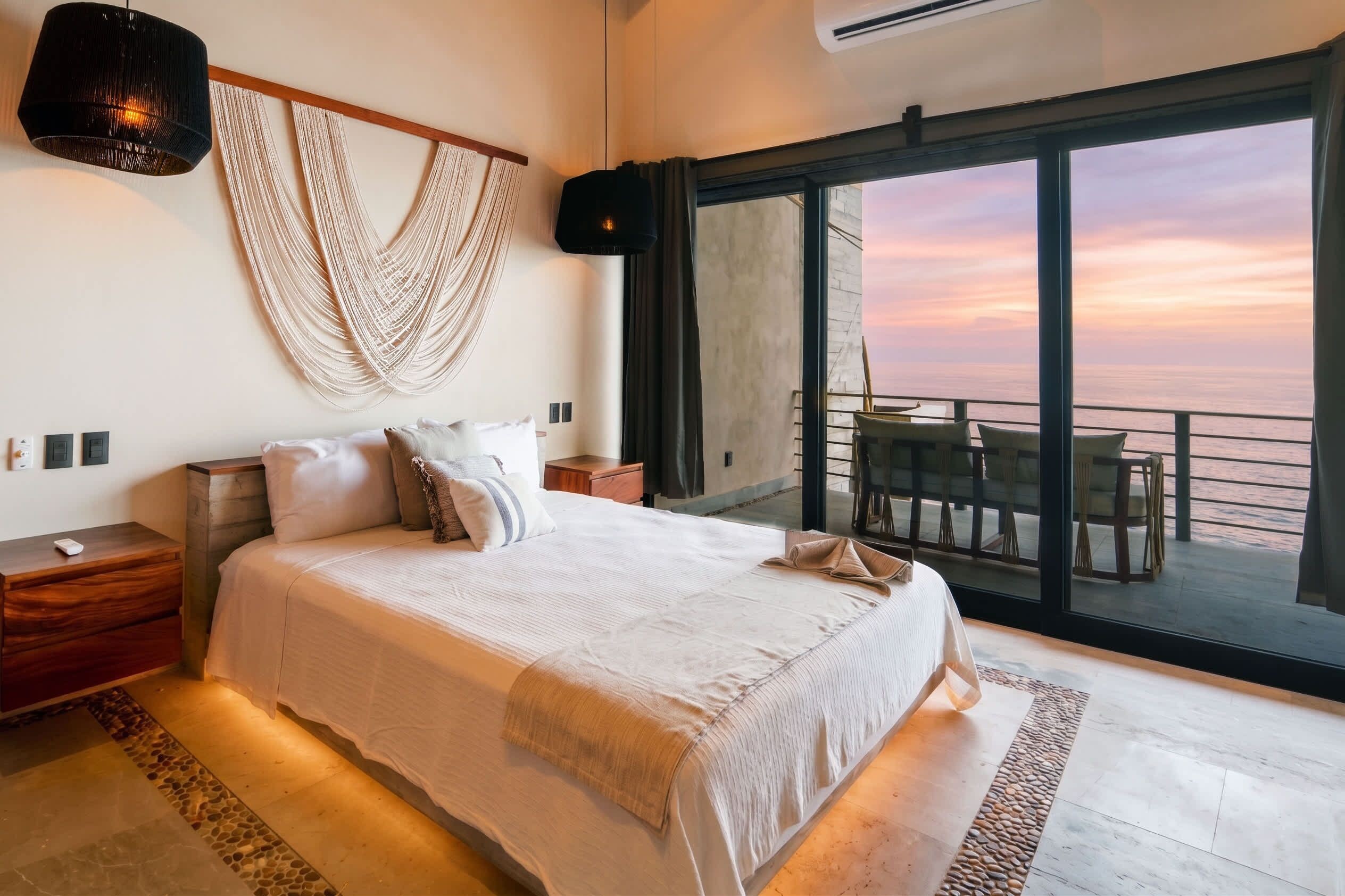 A bedroom with a large bed, a macrame wall hanging, and a view of the sea through a sliding door.