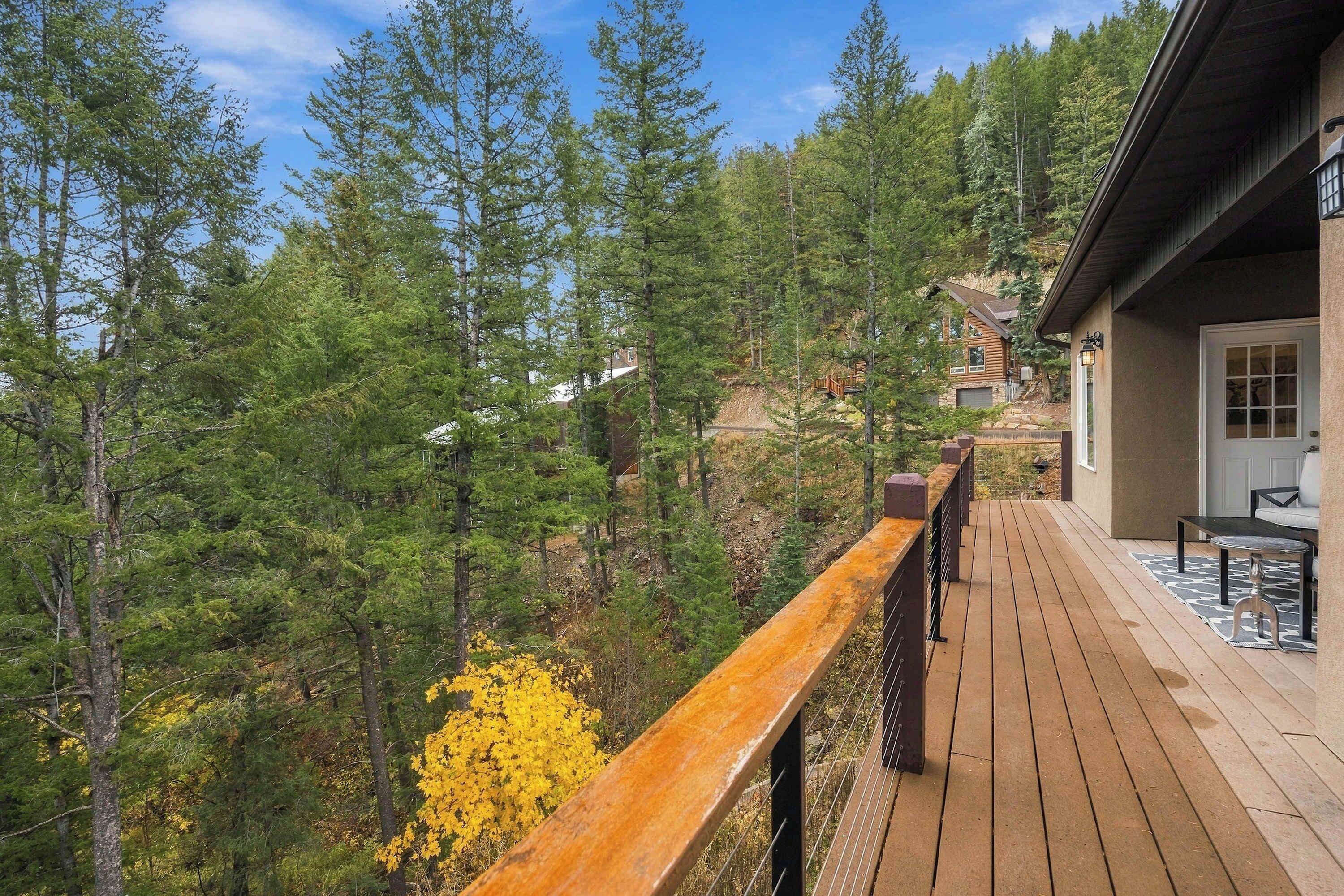 A wooden deck with a railing overlooking a forested area with a house in the background.