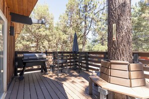 Een houten terras met een barbecue, een bank en een boom.