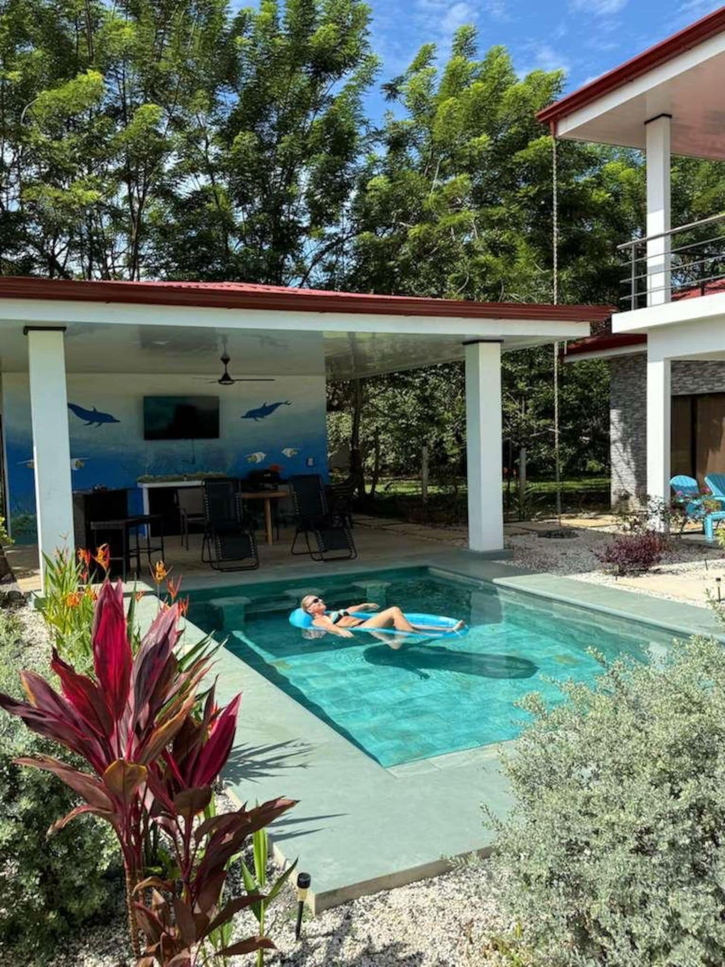 A swimming pool with a person floating on a float, surrounded by plants and a covered area with chairs.