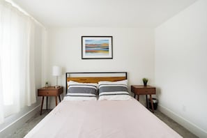 A hotel room with a bed, two bedside tables, a lamp, a framed picture on the wall, and a window with curtains. - Beds for Ten, Bathrooms for a Family • 4BR (Portland)