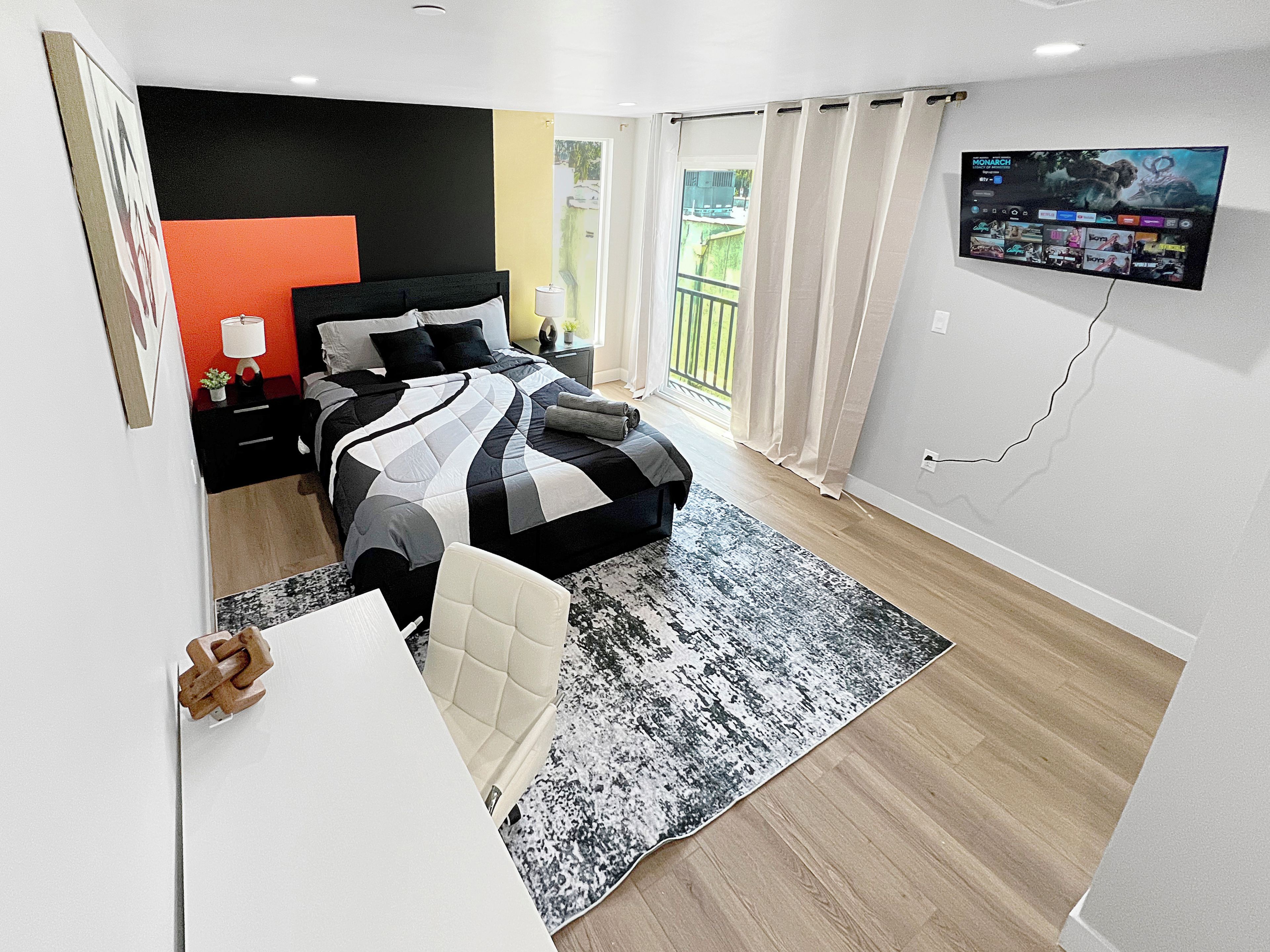 Een moderne slaapkamer met een bed, nachtkastje, lamp en een aan de muur bevestigde televisie.