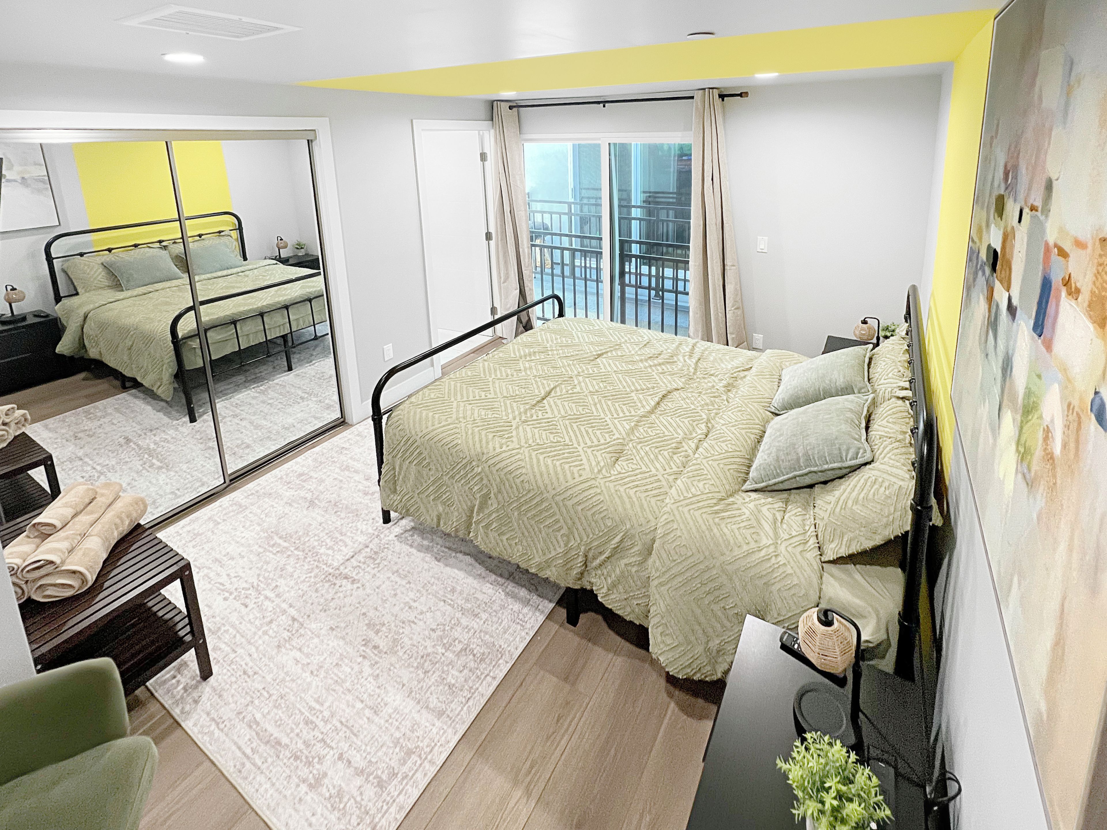 A modern hotel room with a large bed, a desk, a chair, a small table with towels, and a balcony view.