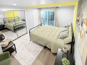 A modern hotel room with a large bed, a desk, a chair, a small table with towels, and a balcony view. - Bedroom with Private Bathroom (Los Angeles)