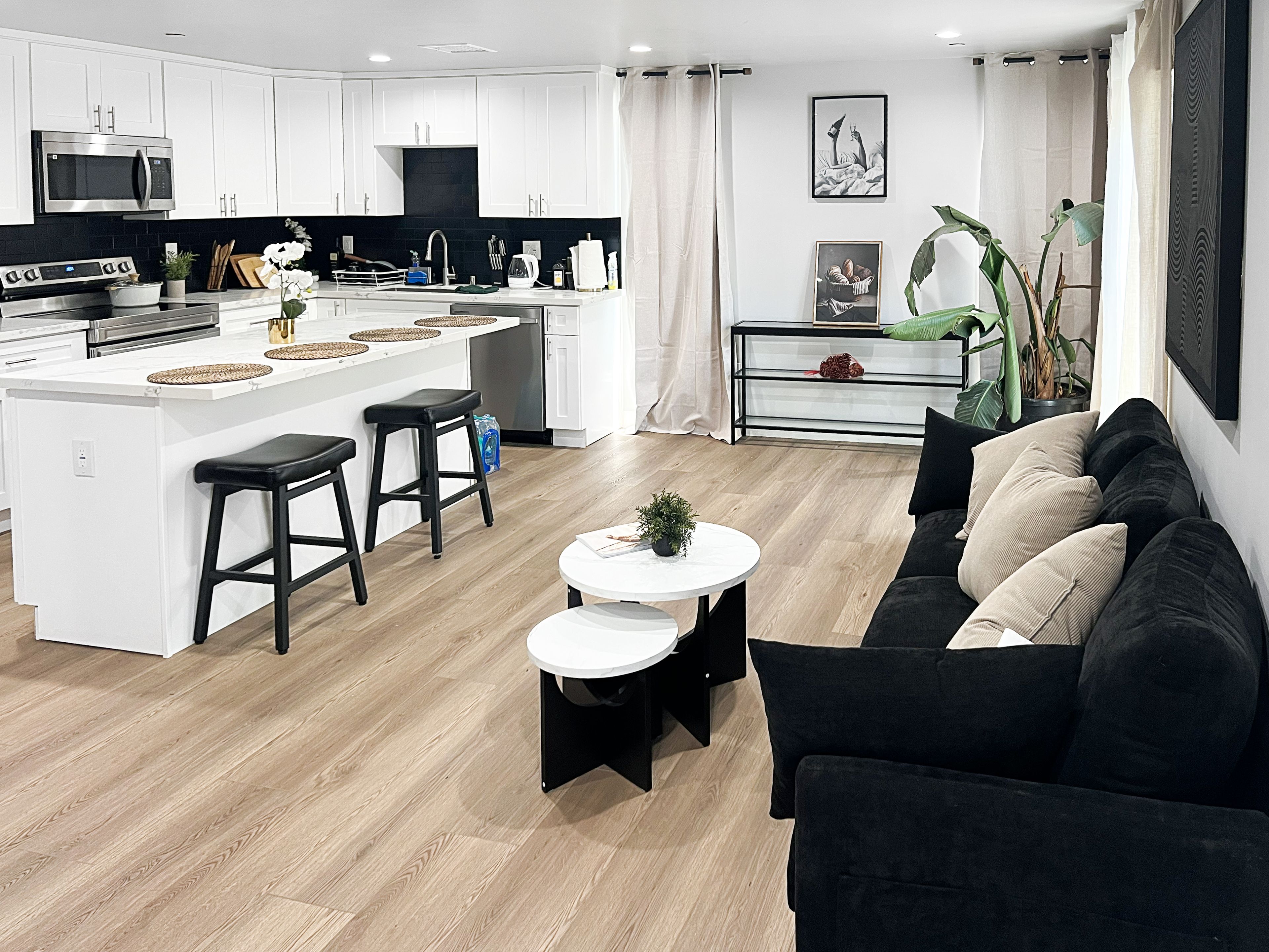 A modern kitchen with white cabinetry, a central island with stools, a dining area with a black sofa and a small round table, and a living room with a black sofa and a coffee table.