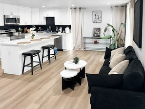 A modern kitchen with white cabinetry, a central island with stools, a dining area with a black sofa and a small round table, and a living room with a black sofa and a coffee table. - Bedroom with Private Bathroom (Los Angeles)