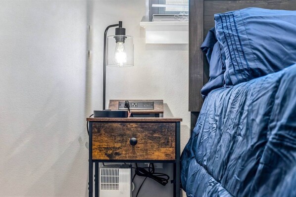 A bedroom with a bed, a bedside table with a lamp, and a power strip. - The Urban Vista Suite *Roof Top Patio Fun!* (Fort Worth)