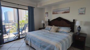 Habitación de hotel con cama, dos mesitas de noche con lámparas, balcón con silla y vista panorámica de la ciudad.
