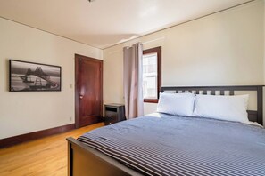 A bedroom with a bed, a nightstand, a door, a window with curtains, and a wall-mounted picture.