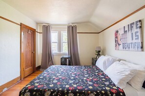 Quarto com cama, janela com cortinas, mesa de cabeceira e abajur, e um pôster na parede.
