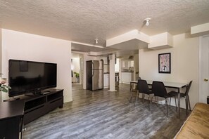 Uma sala de estar moderna com televisão de tela plana, um espaço de jantar com mesa e cadeiras, uma cozinha com armários brancos e uma geladeira.