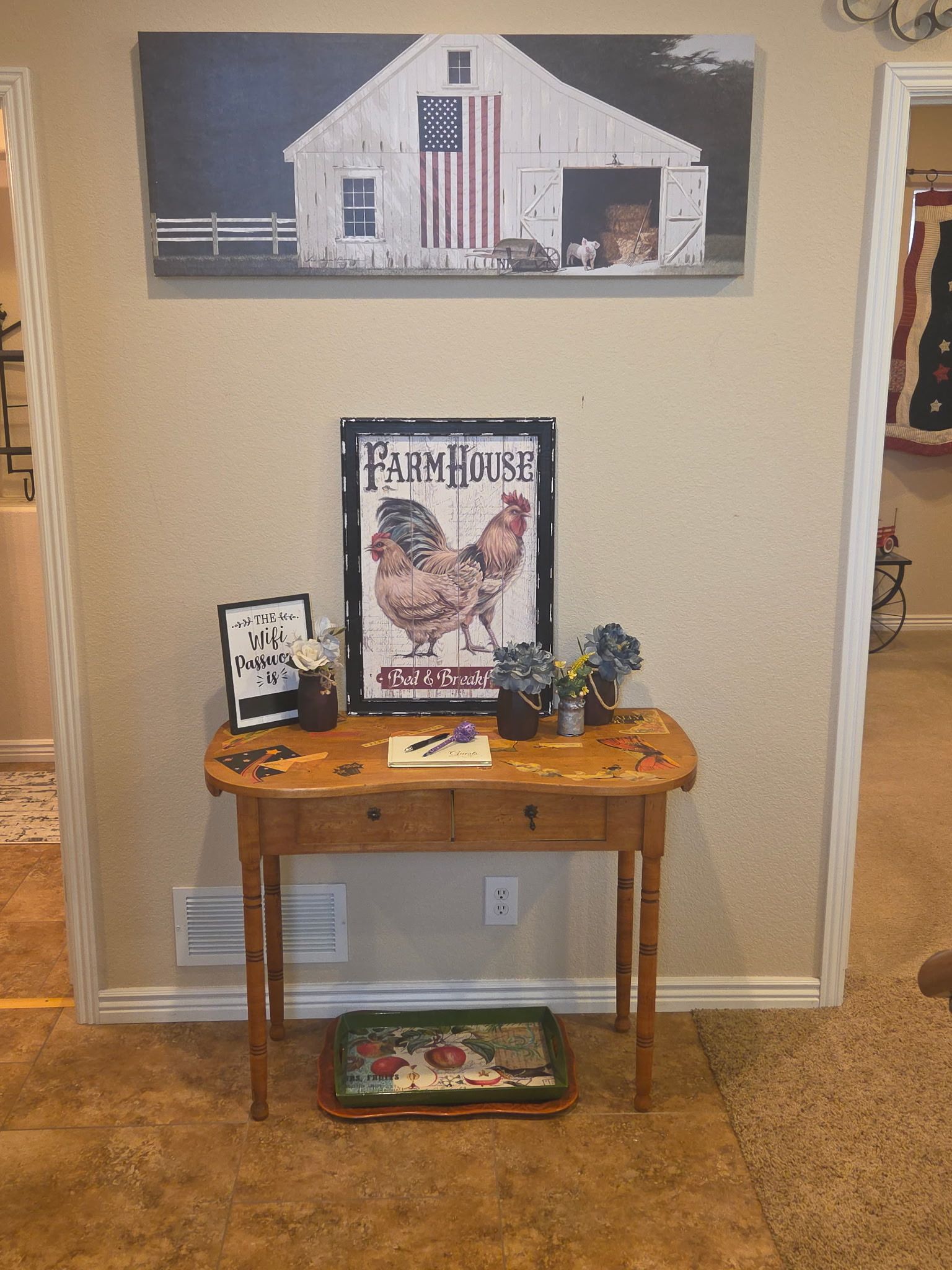 A room with a wall-mounted picture of a barn, a framed sign with a rooster, a small table with decorative items, and a fruit tray on the floor.