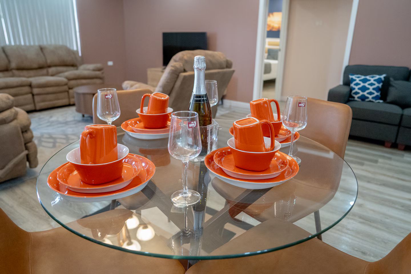 Ein Esstisch, gedeckt mit orangefarbenem Geschirr und Weingläsern, umgeben von Sofas und einem Fernseher in einem Wohnzimmer.
