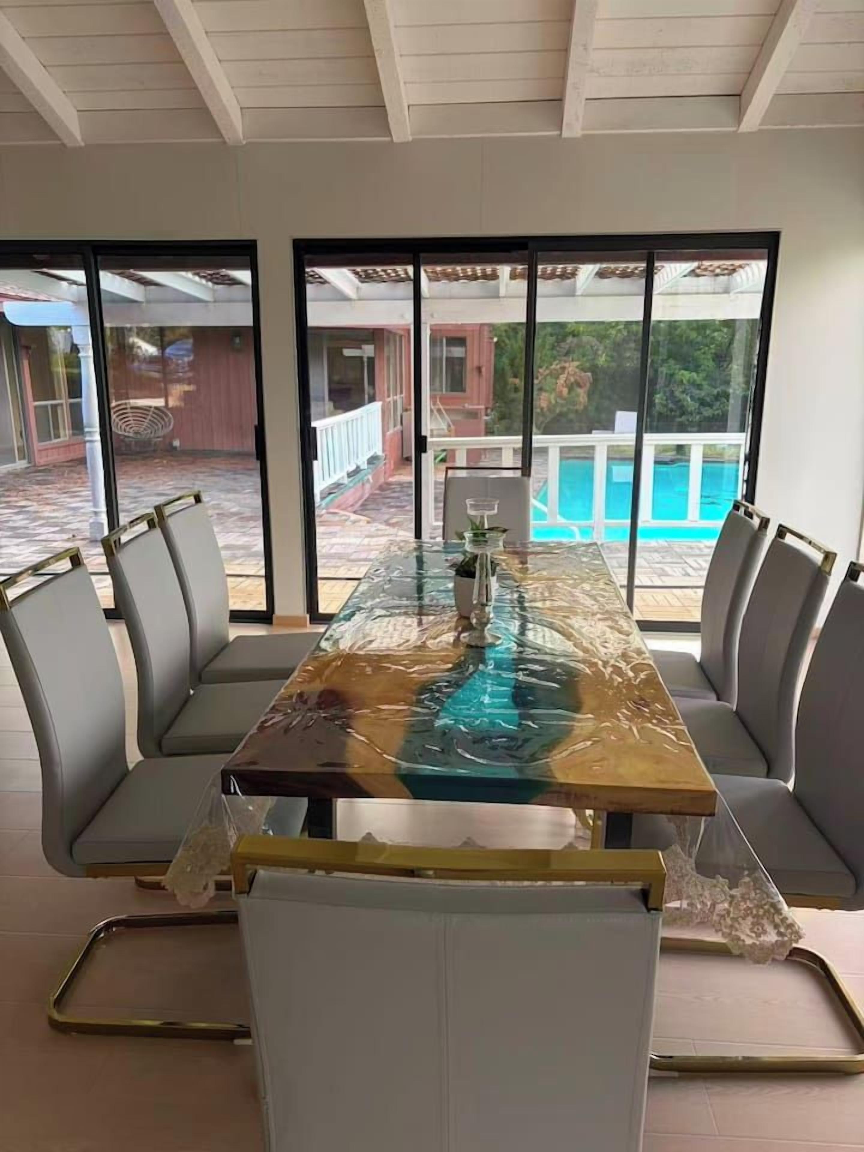 A dining area with a glass-top table, cushioned chairs, and a view of a swimming pool through large windows.