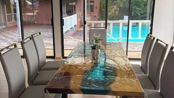 A dining area with a glass-top table, cushioned chairs, and a view of a swimming pool through large windows.