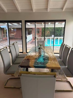Spiseområde med et bord med glasplade, polstrede stole og udsigt til en swimmingpool gennem store vinduer.