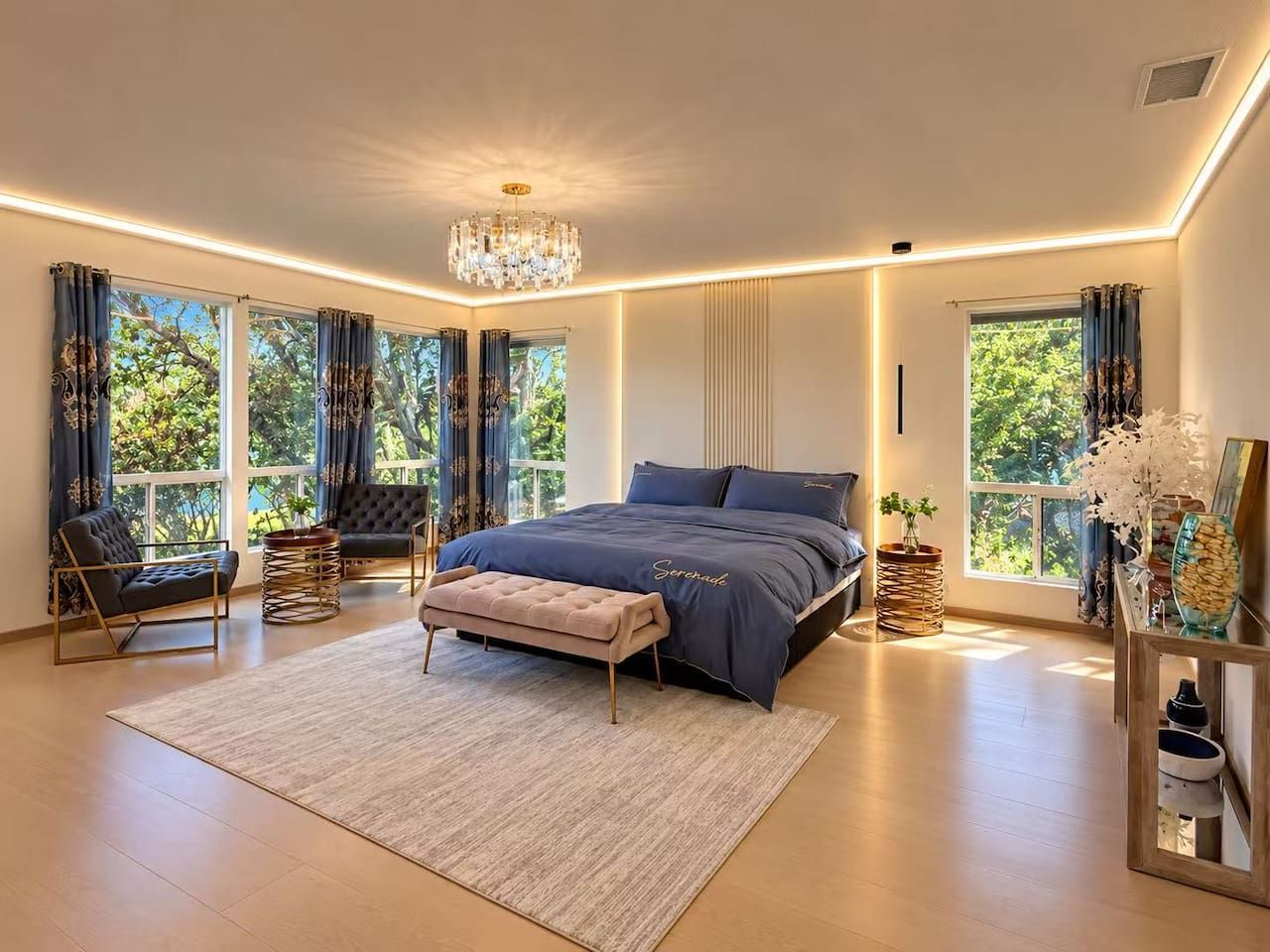 Un dormitorio moderno con una cama grande, una araña y vistas a zonas verdes a través de las ventanas.