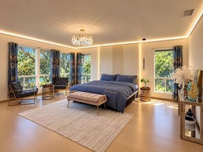 Une chambre moderne avec un grand lit, un lustre et une vue sur la verdure par les fenêtres.