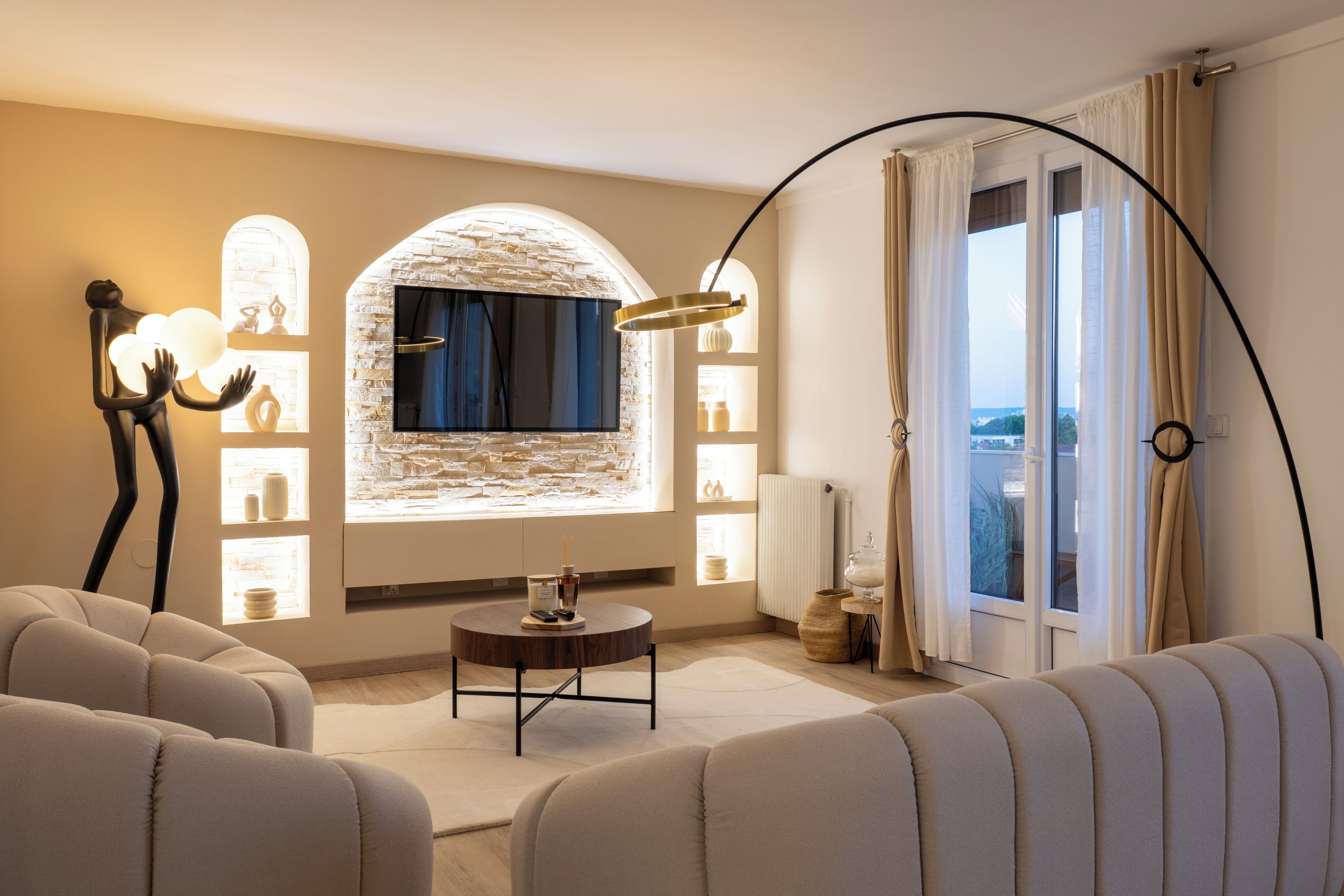 Un salon moderne avec un mur en pierre, un téléviseur à écran plat, une table basse ronde et une grande fenêtre avec des rideaux.