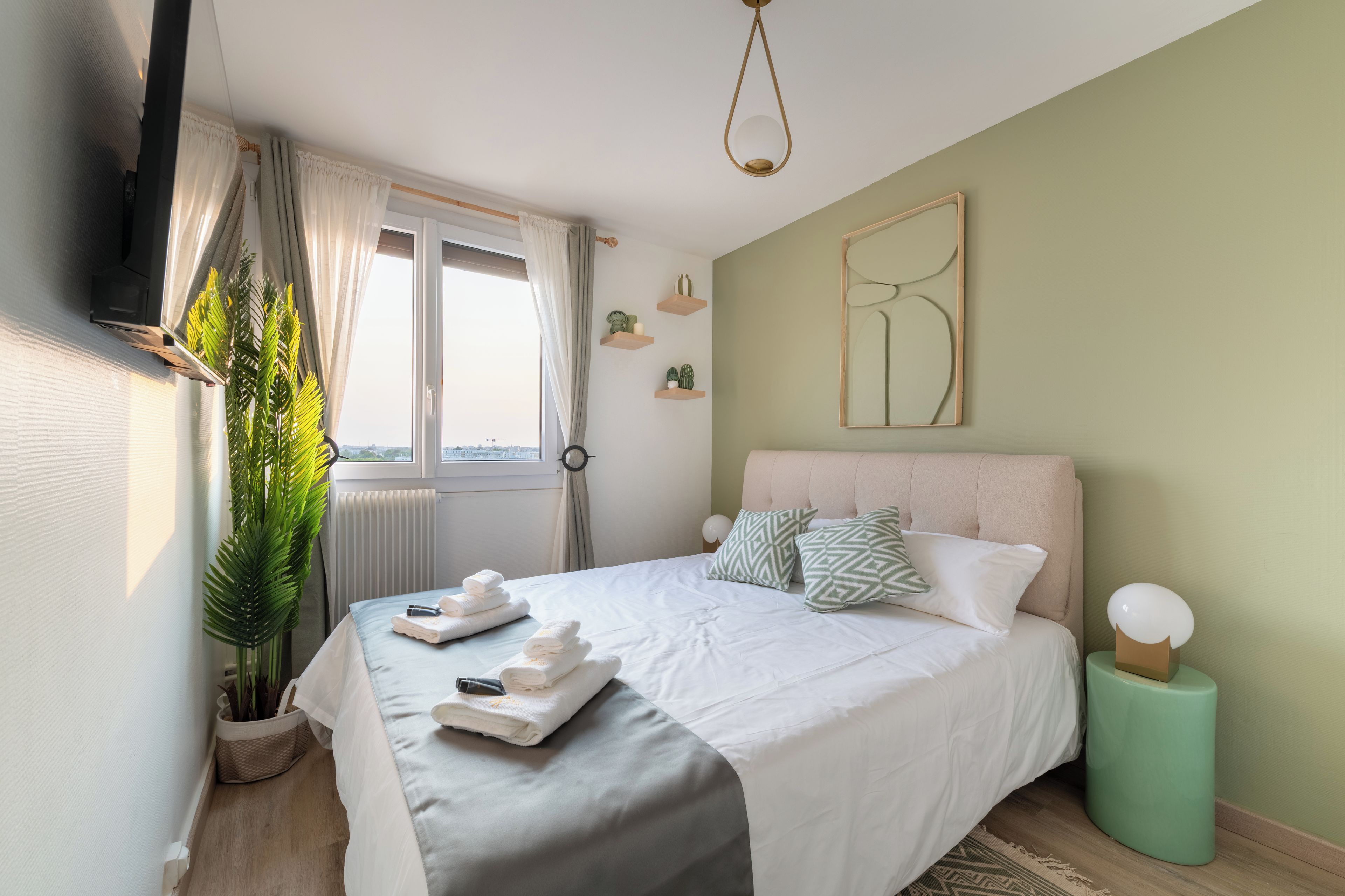 A neatly arranged bedroom with a bed, pillows, a bedside table, a plant, and a window with curtains.