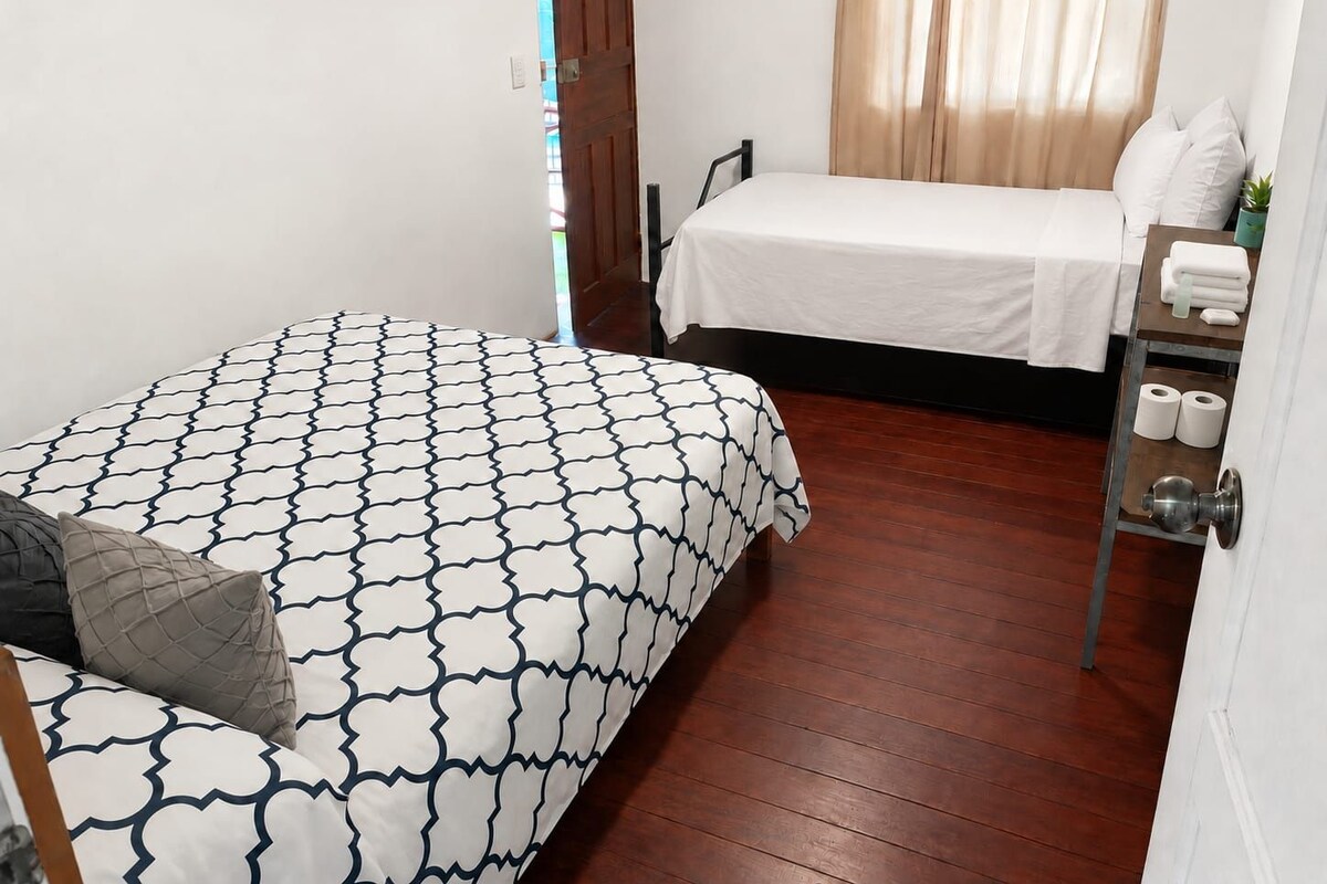 Un dormitorio con dos camas, piso de madera, una puerta y un pequeño estante con artículos de aseo.