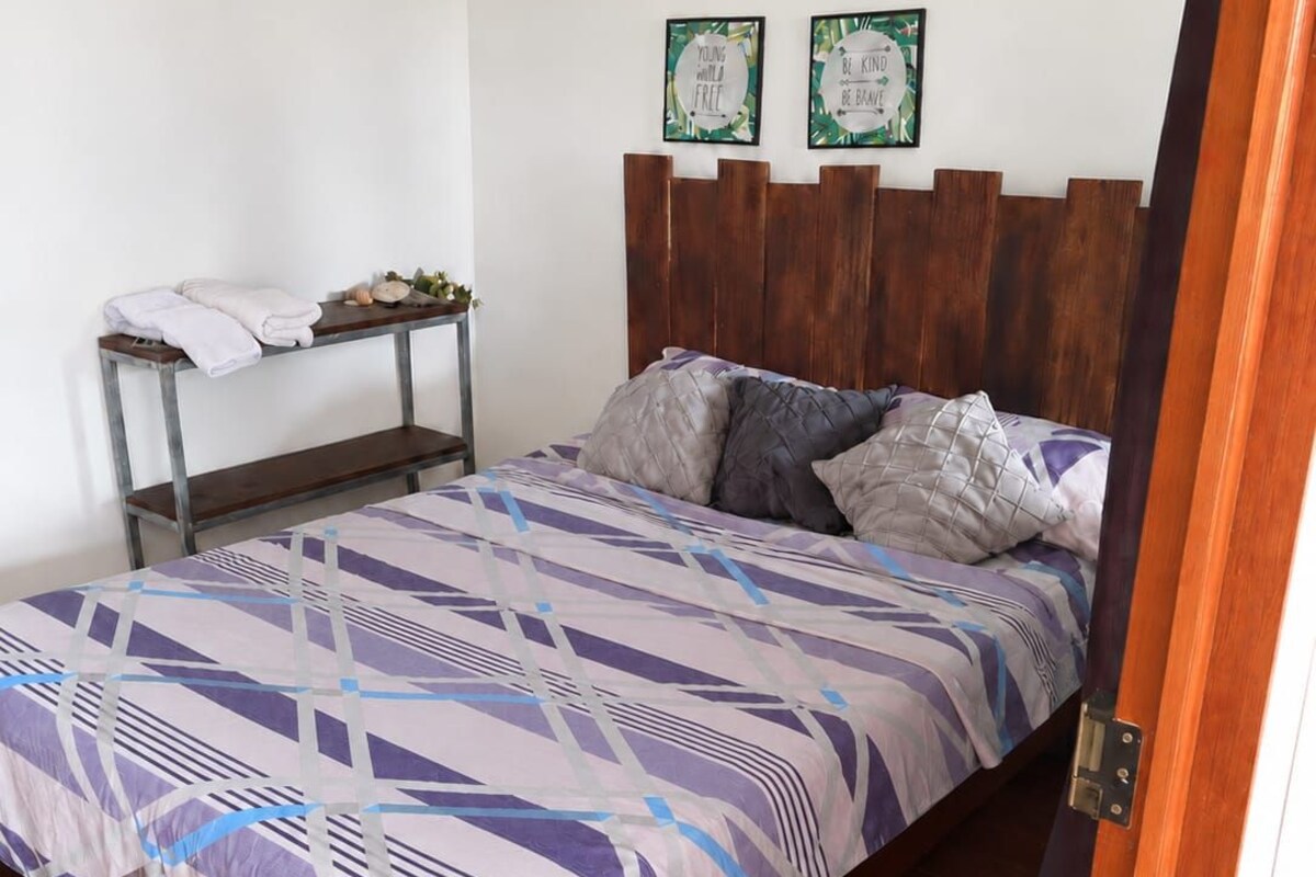 Un dormitorio con cama, una mesita de noche con toallas, aire acondicionado de pared y dos carteles decorativos con frases inspiradoras.