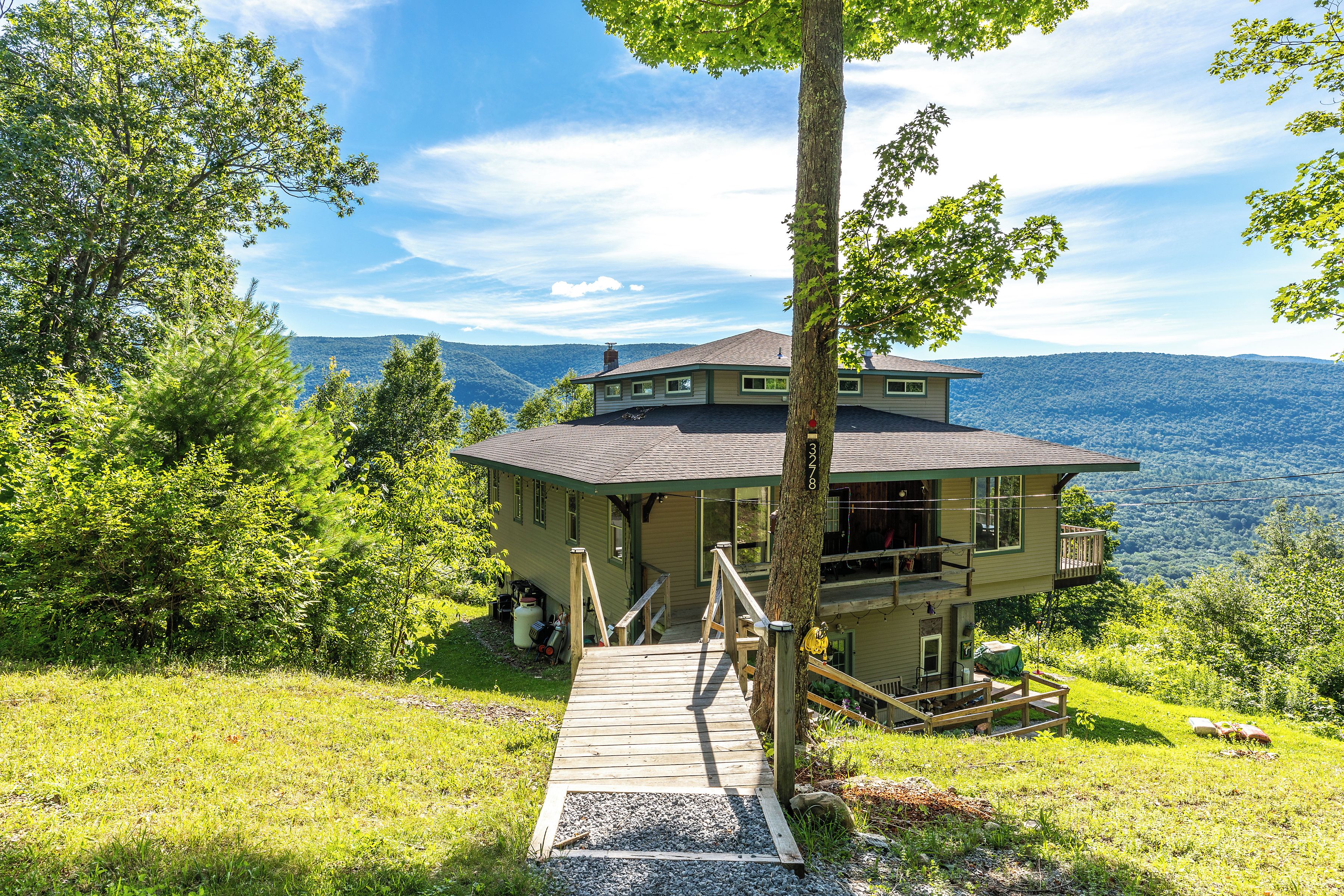 Ein zweistöckiges Haus mit einer Holzterrasse, die zu einer Haustür führt, umgeben von üppiger Vegetation und einer Bergkulisse.
