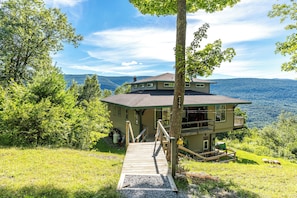 A two-story house with a wooden deck leading to a front door, surrounded by lush greenery and a mountainous backdrop. - Wonderful hilltop house with breathtaking views (Dorset)