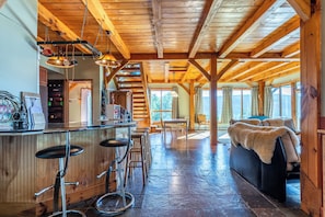 A rustic interior with a wooden ceiling, a bar area with stools, a sofa, and a dining table.