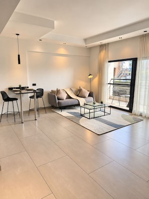 A modern living room with a sofa, coffee table, dining table, chairs, and a large window with curtains. - Sky Suites Quattro (Accra)