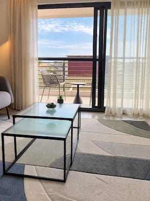 A room with a glass table, a chair, a small potted plant, and a view of the sea through a balcony with a railing. - Sky Suites Quattro (Accra)