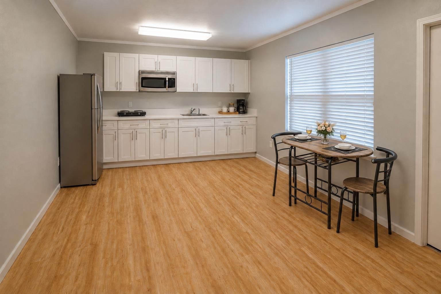 A compact kitchen with white cabinets, a stainless steel refrigerator, a microwave, and a coffee maker. There is a dining table with two chairs and a window with blinds.
