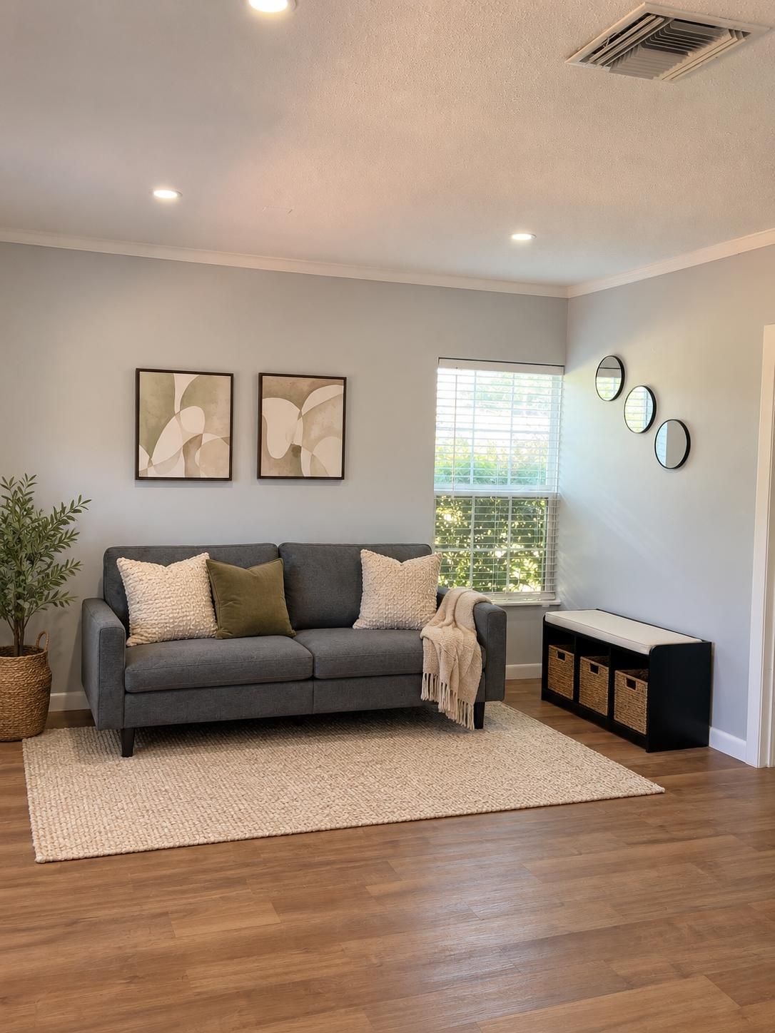 A modern living room with a grey sofa, two abstract paintings, a window with blinds, and a bench with baskets.