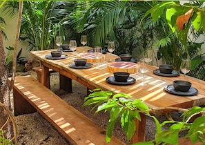 Un espace de restauration en plein air avec une table en bois dressée pour six personnes, entourée d’une végétation luxuriante.