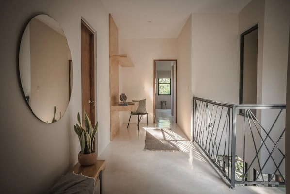 Un couloir avec un miroir rond, une plante en pot, une chaise et un escalier doté d’une rampe en métal.