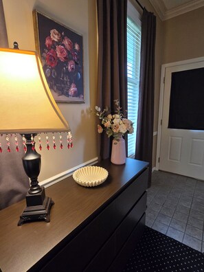 Una habitación con escritorio, lámpara, florero con flores, un cuadro enmarco y una puerta.