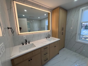 A modern bathroom with a large mirror, two sinks, wooden cabinets, and a window.