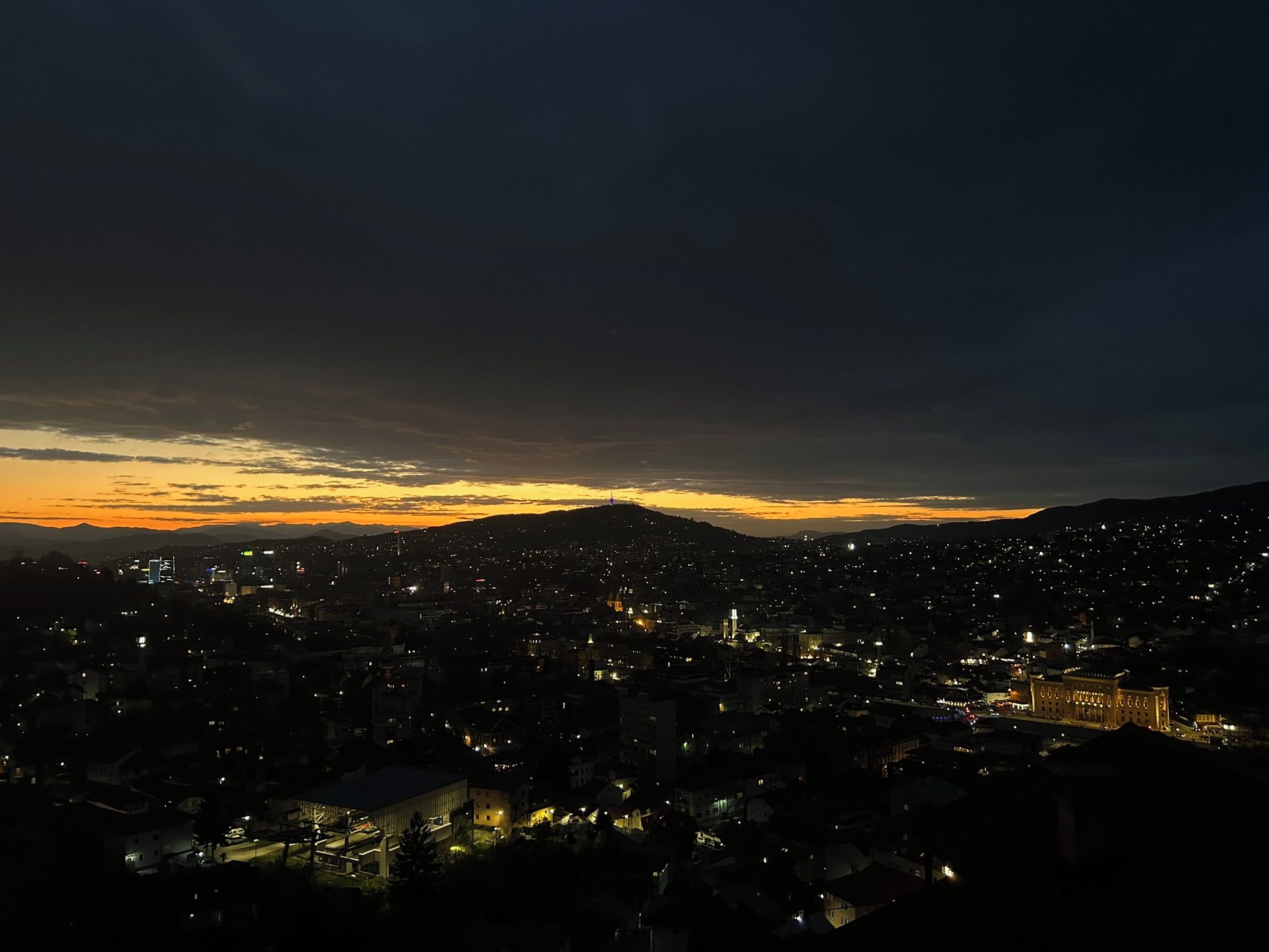 Paisagem urbana ao anoitecer, com edifícios iluminados e uma cordilheira ao fundo.