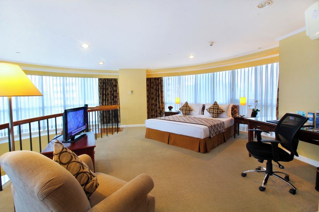 Habitación de hotel con cama, escritorio, silla, televisor y ventanas amplias.