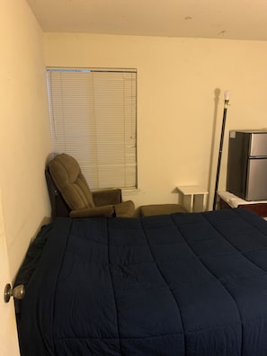 A bedroom with a bed, a chair, a small table, a lamp, and a refrigerator.