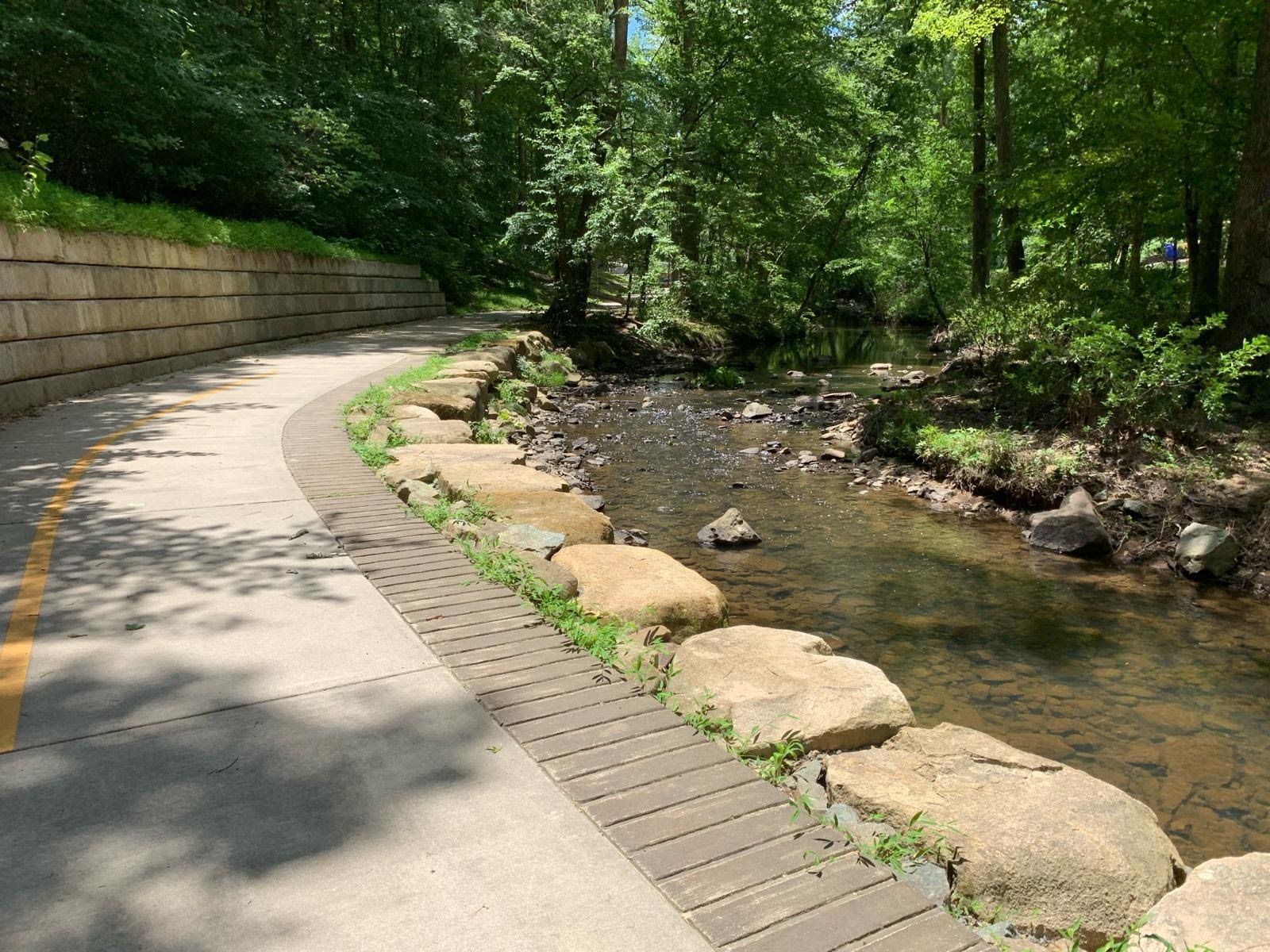 A curved pathway with a concrete edge and a wooden boardwalk alongside a clear stream with rocks.
