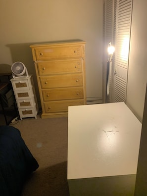 A room with a white table, a wooden dresser, a white nightstand with a fan, and a wall-mounted heater.