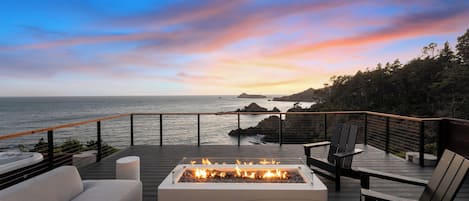 A deck with a fire feature, seating, and a view of the ocean at sunset.
