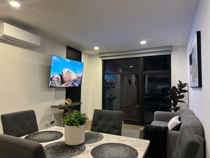 A modern living room with a flat-screen TV mounted on the wall, a dining area with a table and chairs, and a sliding glass door leading to a balcony. - New Listing (Tijuana)